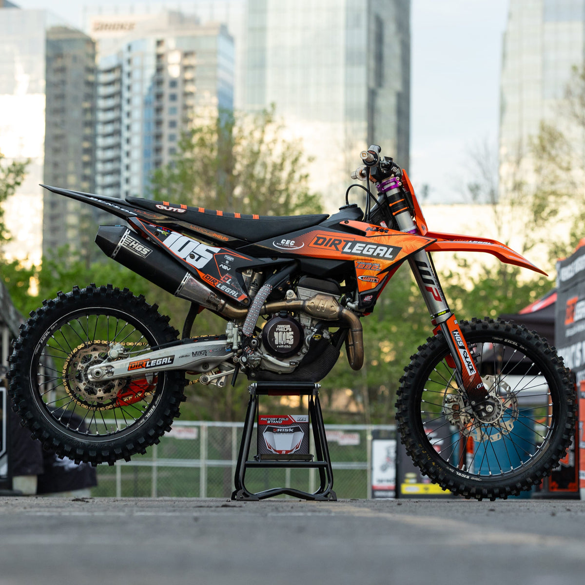 Moto da cross arancione e nera su una Risk Racing F.F.S. stare con il paesaggio urbano sullo sfondo