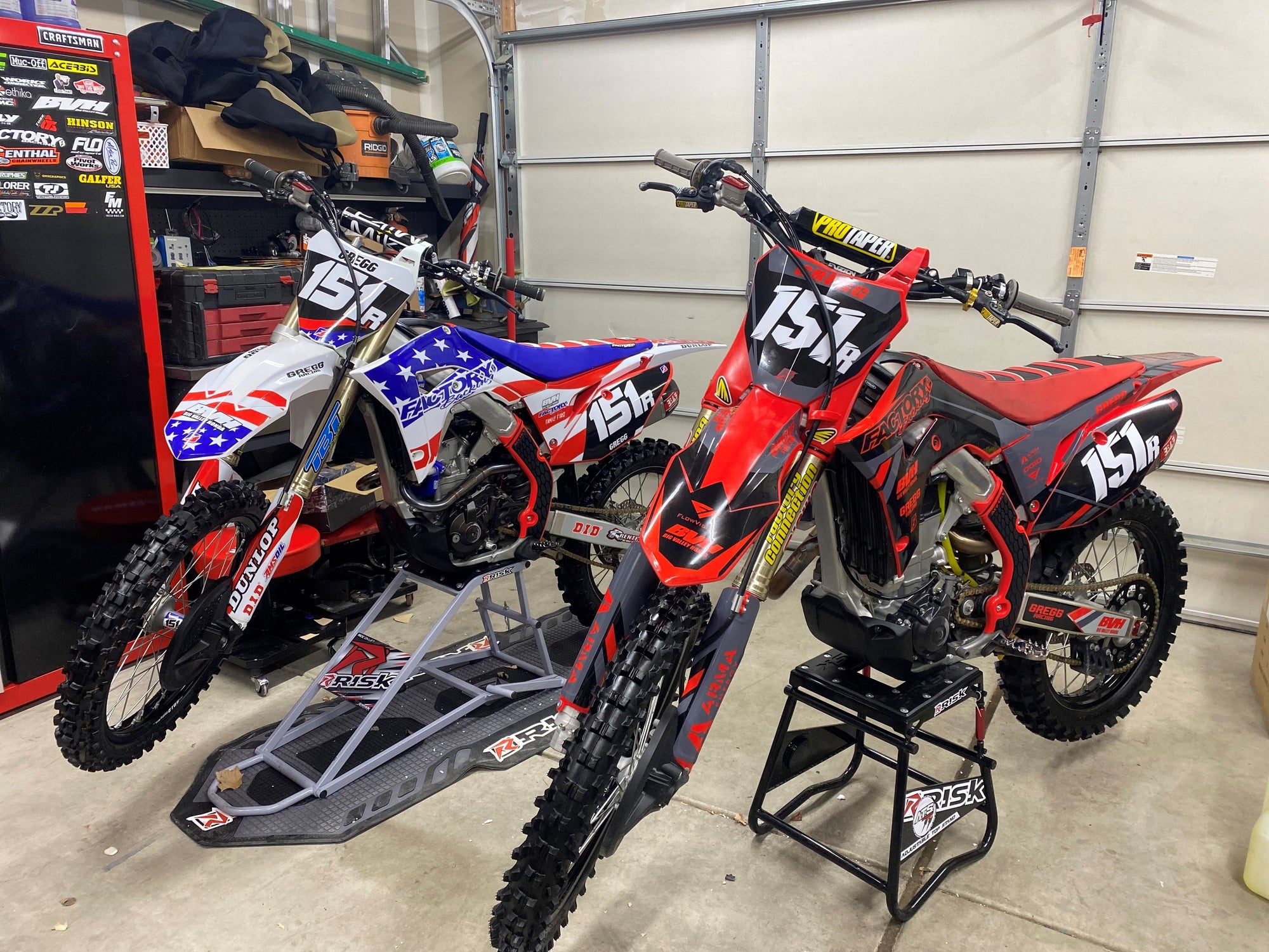 two motocross dirt bikes in a garage. One is sitting on an RR1 Ride on Lift and Factory pit mat and the other is on an ATS adjustable top stand. MX equipment by Risk Racing.