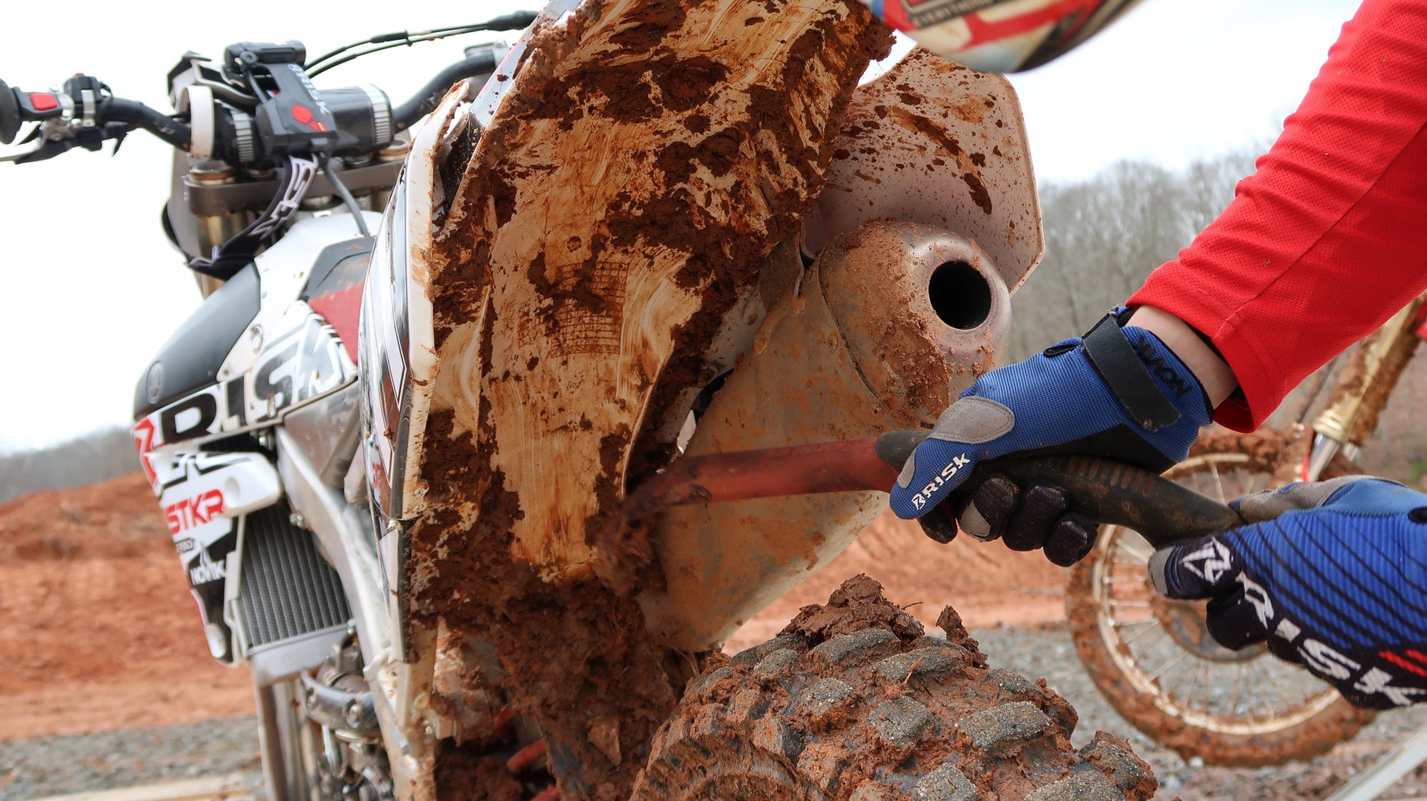 close up of someone scraping the mud from underneath their rear dirt bike fender using a mud axe from Risk Racing